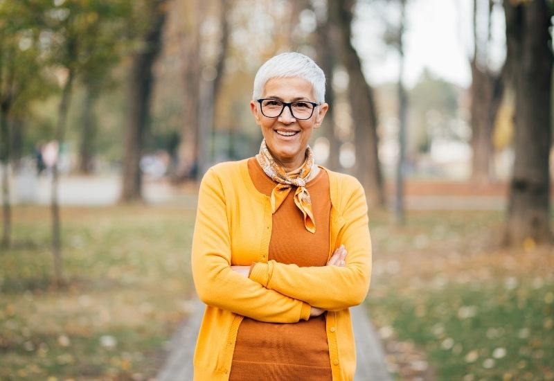 Older woman standing in woods, smiling with arms crossed
